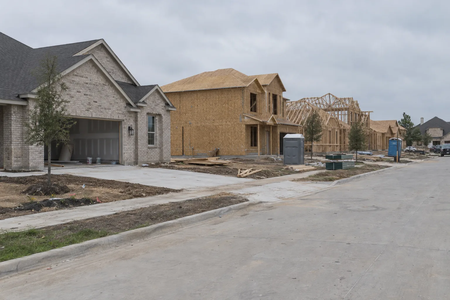 A suburban Dallas street showing several new construction homes at different stages of building, from bare wood framing to nearly complete brick exteriors, with lumber stacks and construction materials on flat north Texas lots under a hazy overcast sky