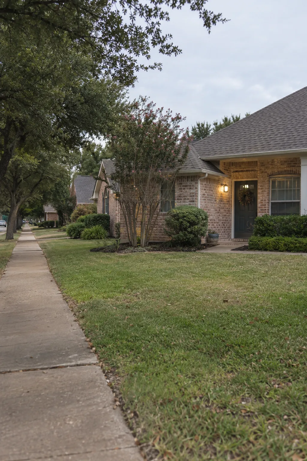 A quiet suburban street lined with single-story brick ranch homes in the Dallas-Fort Worth area, bathed in warm golden-hour light, with crepe myrtles, live oaks, and front porches visible along an empty sidewalk
