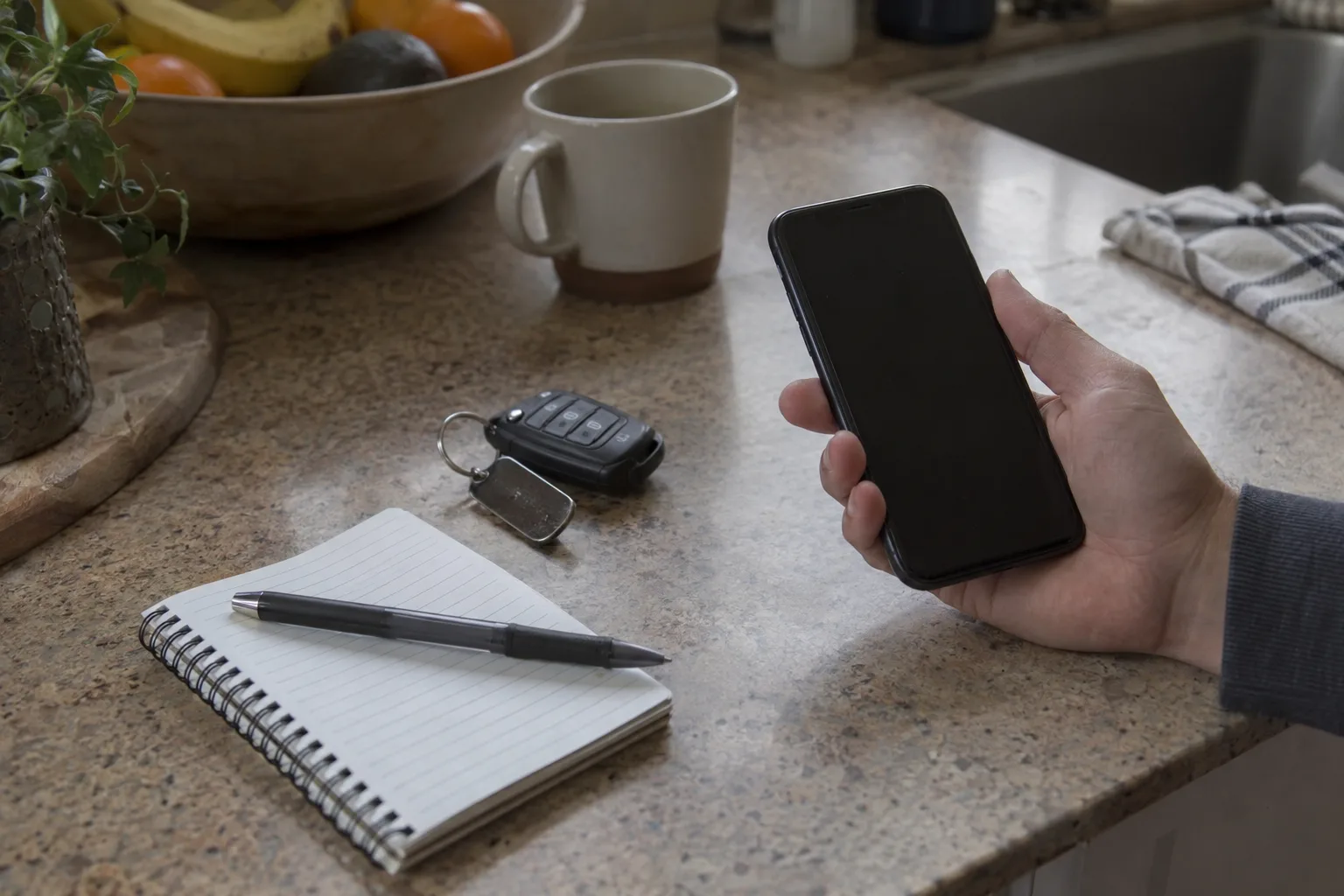 A hand holding a smartphone to the ear beside a car key fob and a blank spiral notepad with a pen on a sunlit kitchen counter