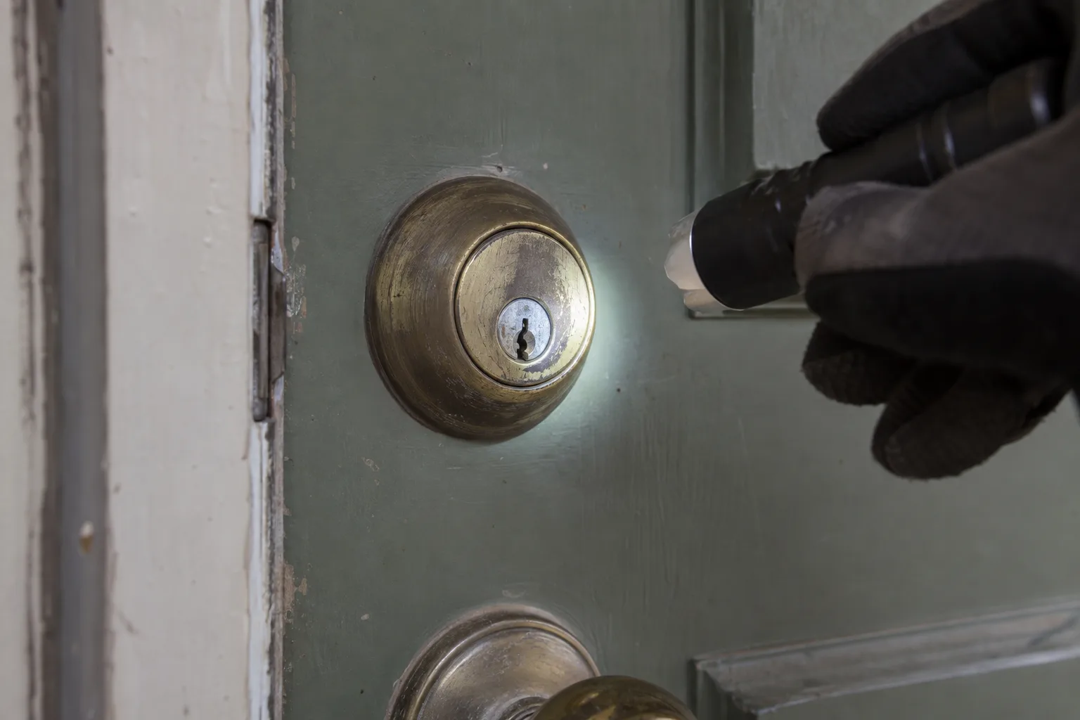 A gloved hand holding a small flashlight shines light into the keyway of a worn brass deadbolt mounted on a sage-green residential front door, revealing scratches and visible wear around the cylinder face