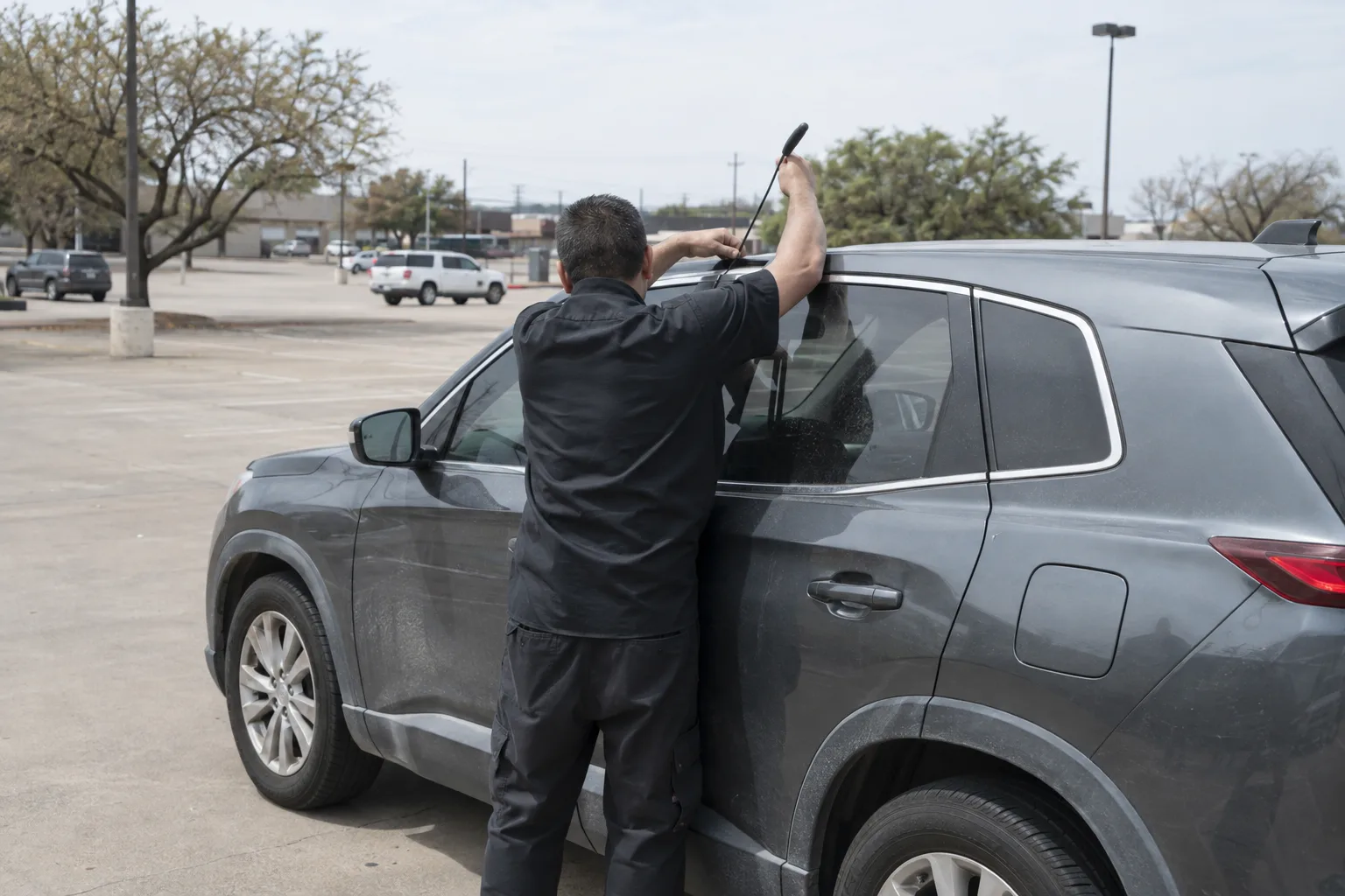 A technician facing away from the camera works at the driver's door of a grey SUV in a sunny parking lot, threading a long slender tool into the top of the door frame.
